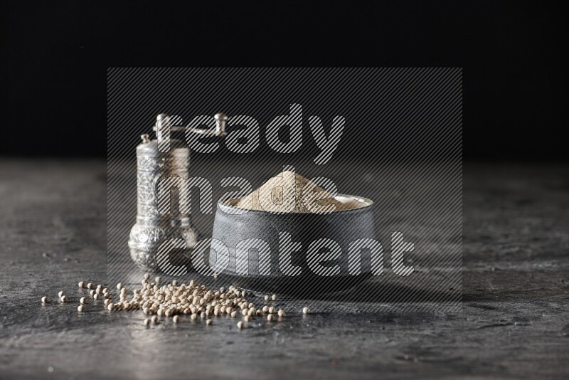 وعاء فخار أسود مملوء ببودرة الفلفل الأبيض مع حبات الفلفل ومطحنة الفلفل المعدنية على أرضية سوداء