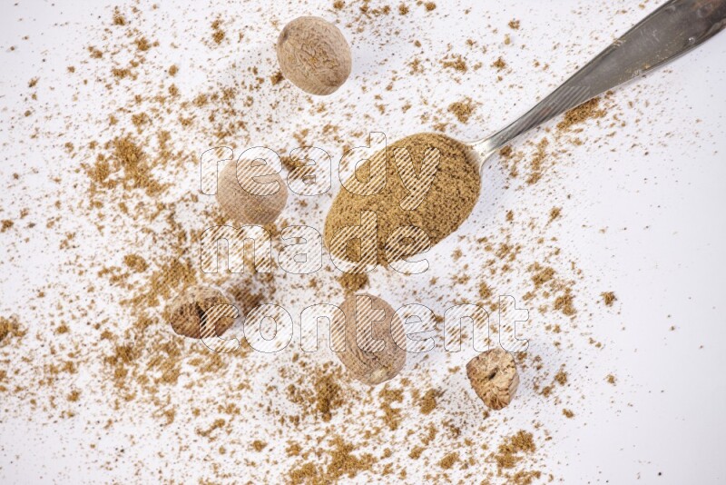 A metal spoon full of nutmeg powder with some of the powder and seeds beside it on a white flooring