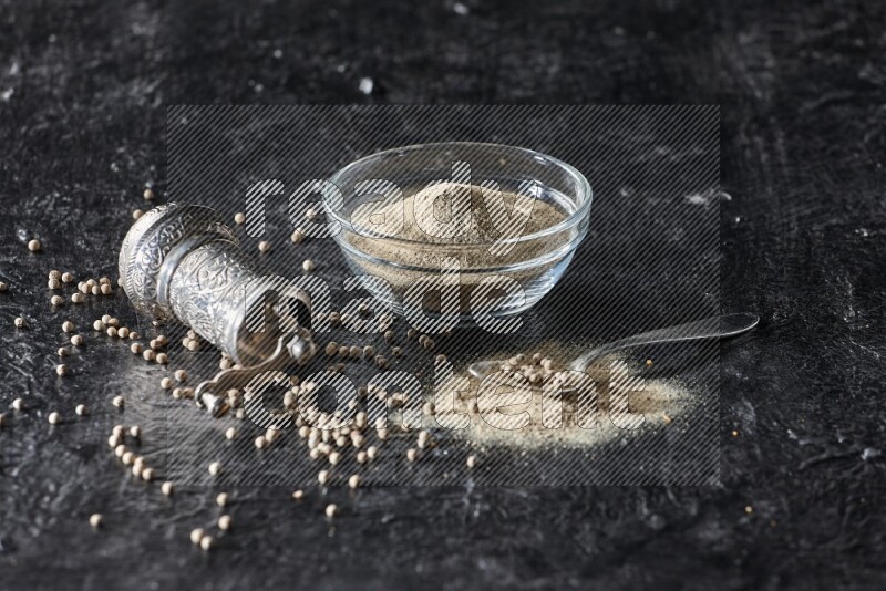 A glass bowl full of white pepper powder with pepper beads, a metal grinder and a metal spoon on textured black flooring