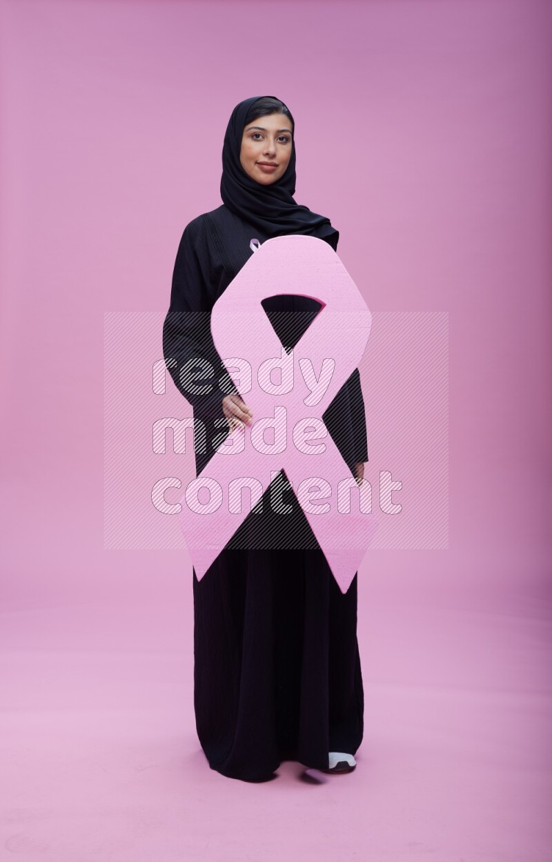 Saudi woman wearing pink ribbon on abaya standing holding awareness ribbon on plain pink background