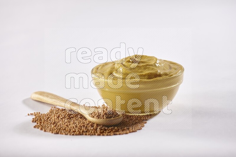 A glass bowl full of mustard paste with some of mustard seeds underneath it and a wooden spoon on white flooring