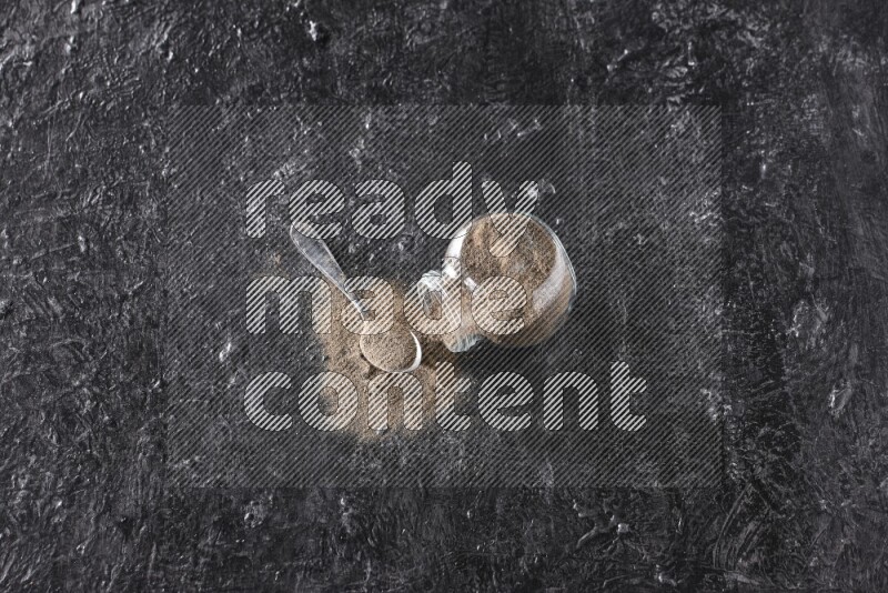 بودرة الفلفل الأسود في وعاء زجاجي مقلوب مع ملعقة معدنية مليئة بالبودرة علي خلفية سوداء