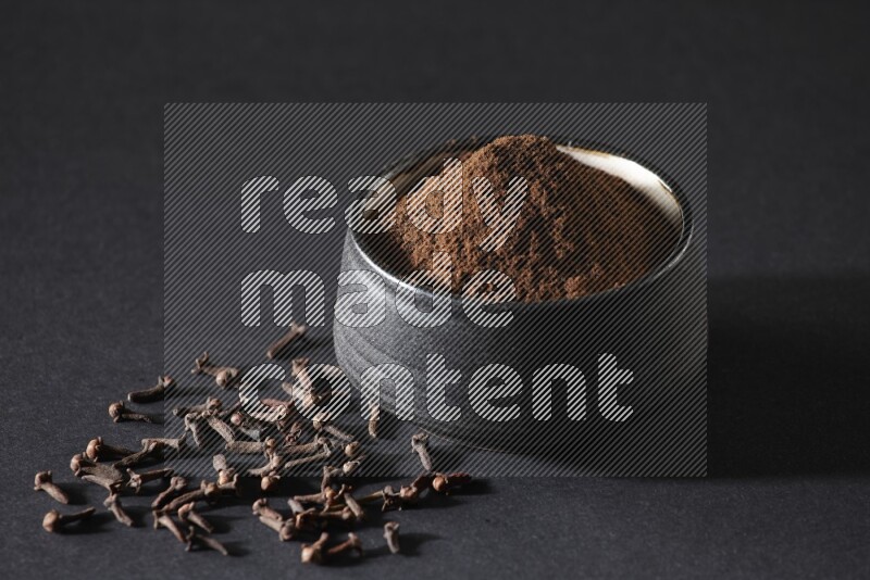 A black pottery bowl full of cloves powder and cloves whole grains on a black flooring