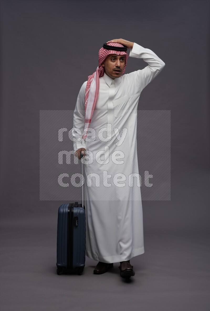 Saudi man wearing thob and shomag standing holding bag on gray background