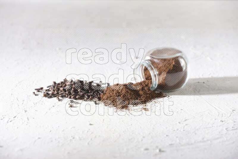 A glass spice jar full of cloves powder flipped and powder came out of it with cloves spread on textured white flooring