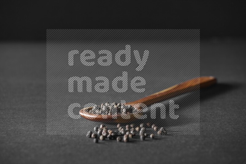A wooden ladle full of black pepper beads on a black flooring