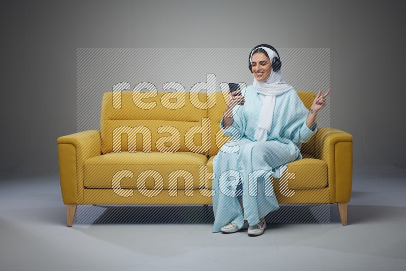 A Saudi woman wearing a light blue Abaya and a white head scarf sitting on a yellow sofa and holding an iPad while wearing headphone eye level on a grey background