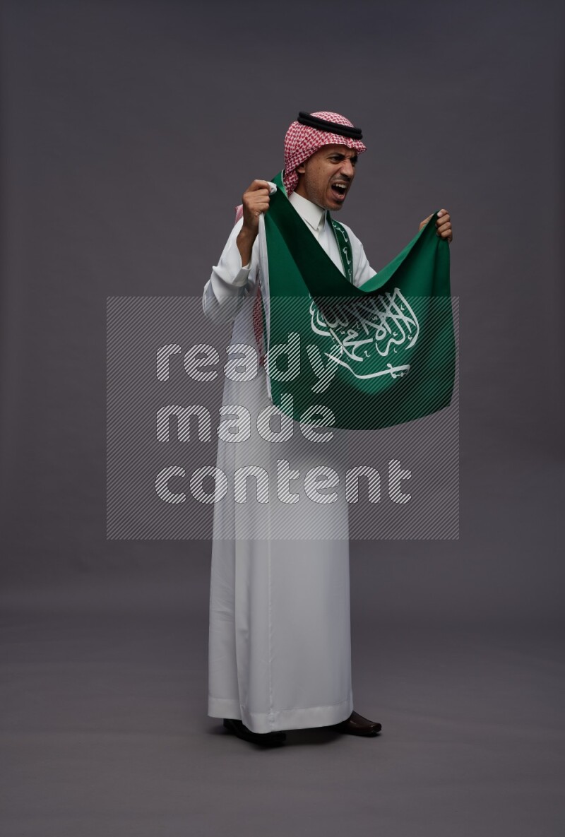 Saudi man wearing thob and shomag standing holding Saudi flag on gray background
