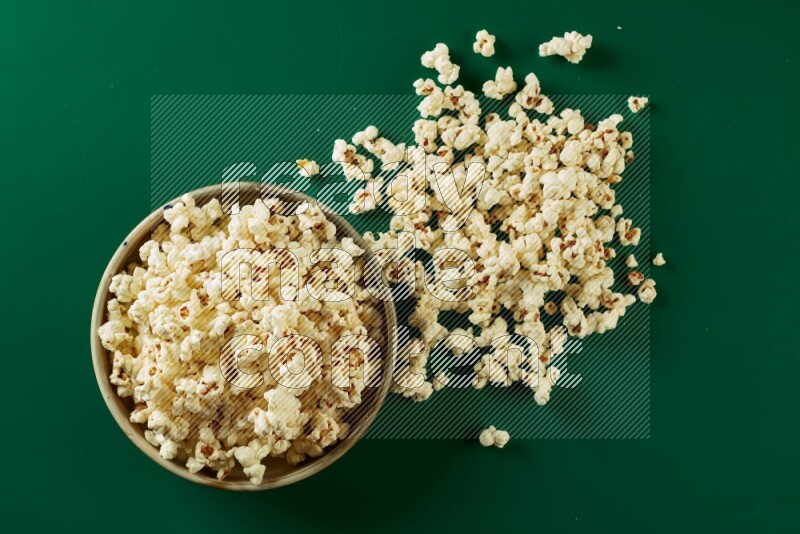 A beige ceramic bowl full of popcorn with popcorn beside it on a green background in a top view shot