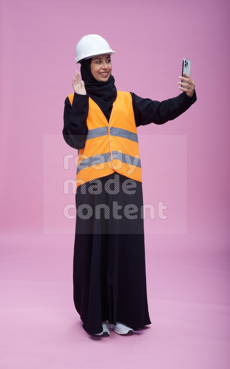 Saudi woman wearing Abaya with engineer vest and helmet standing taking selfie on pink background