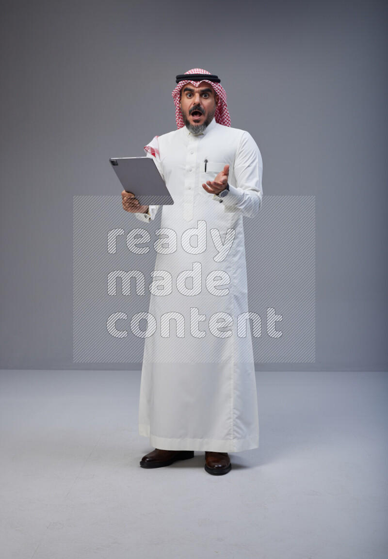 Saudi man Wearing Thob and red Shomag standing working on tablet on Gray background