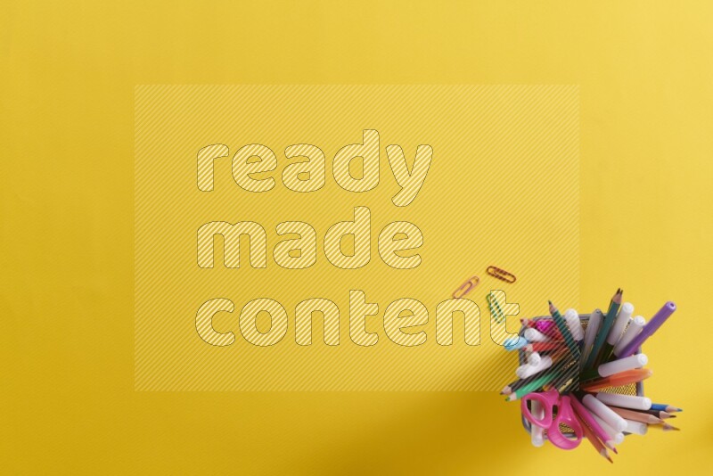 Pens, pencils and school supplies in a metal pen holder on yellow background