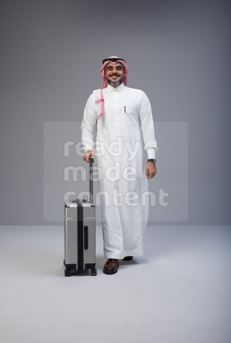 Saudi man wearing Thob and red Shomag standing holding Travel bag on Gray background