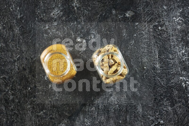 2 glass spice jars full of turmeric powder and dried whole fingers on textured black flooring