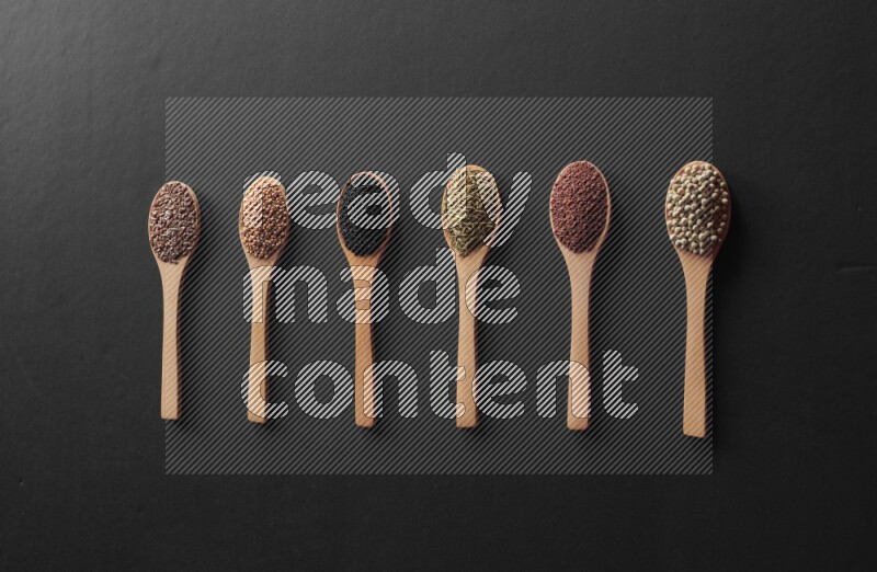 wooden spoons filled with white peppers, mustard seeds, black seeds, garden cress, cumin and flax on black flooring