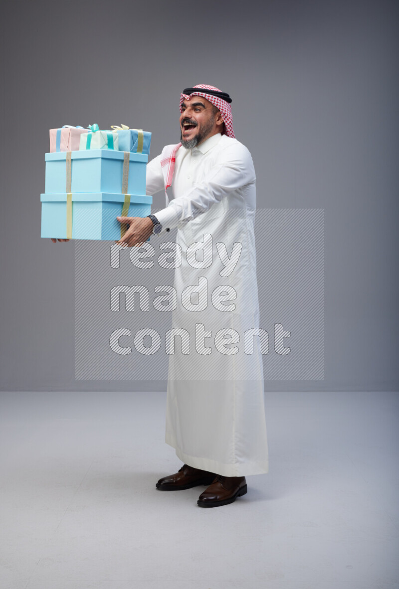 Saudi man Wearing Thob and red Shomag standing holding gift box on Gray background