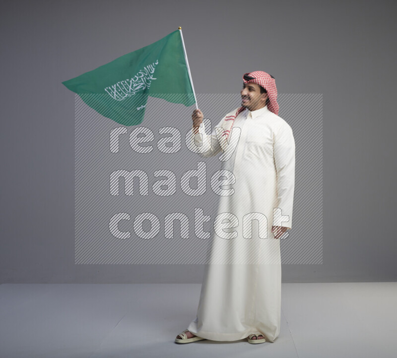 A Saudi man standing wearing thob and red shomag raising big Saudi flag on gray background