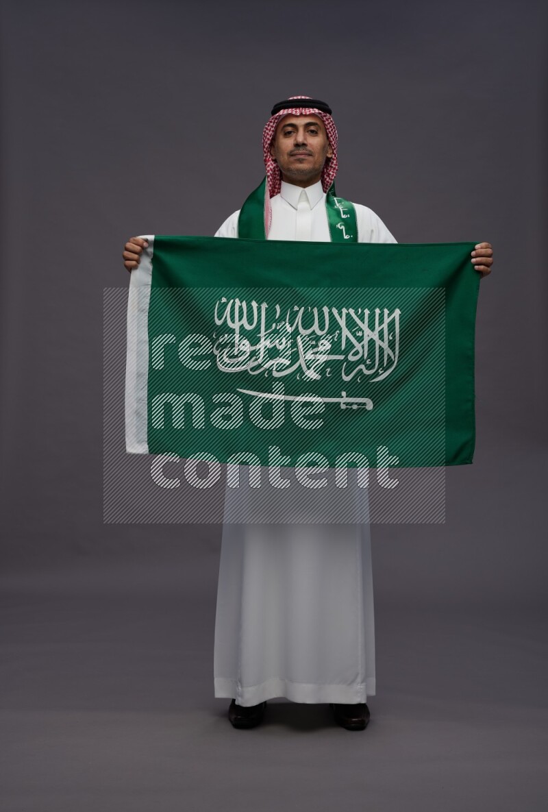 Saudi man wearing thob and shomag standing holding Saudi flag on gray background