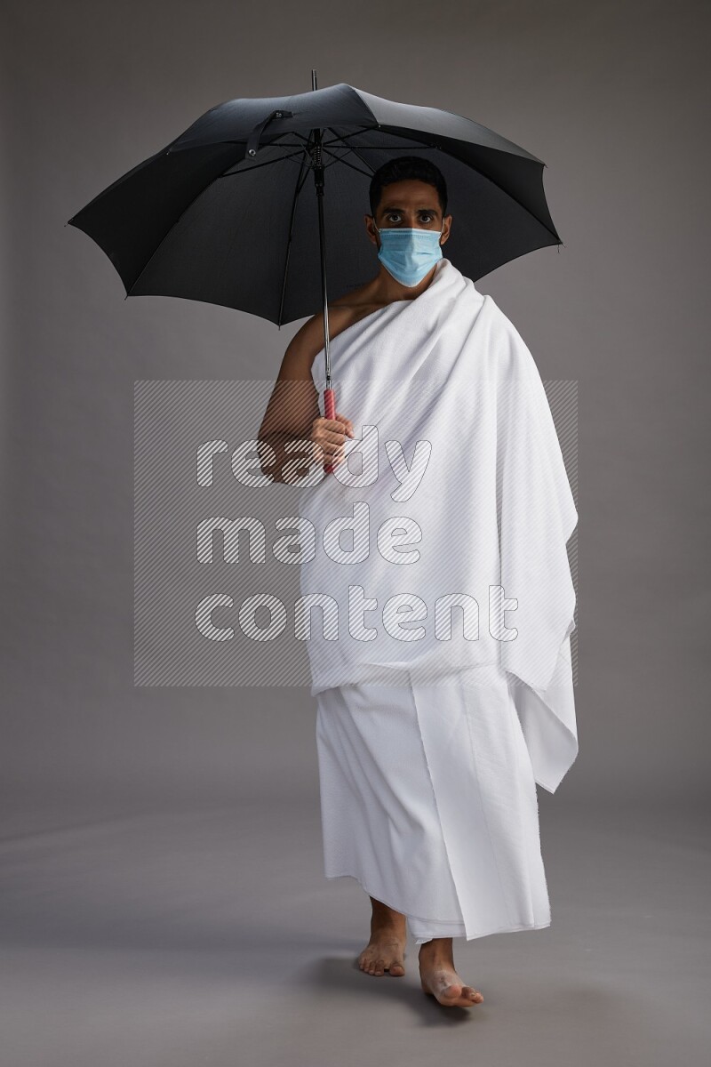 A man wearing Ehram and mask Standing holding umbrella on gray background on gray background