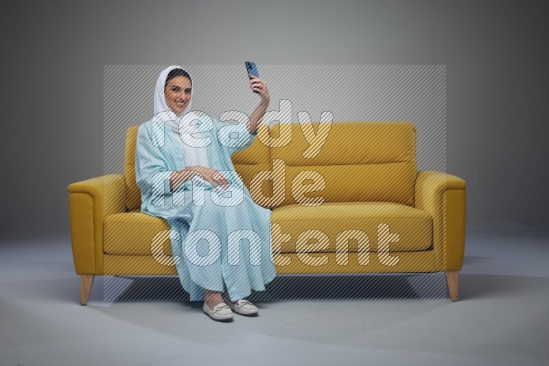 A Saudi woman wearing a light blue Abaya and white head scarf sitting on a yellow sofa and taking selfies with her phone eye level on a grey background