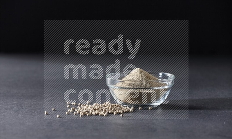 A glass bowl full of white pepper powder with white pepper beads on black flooring