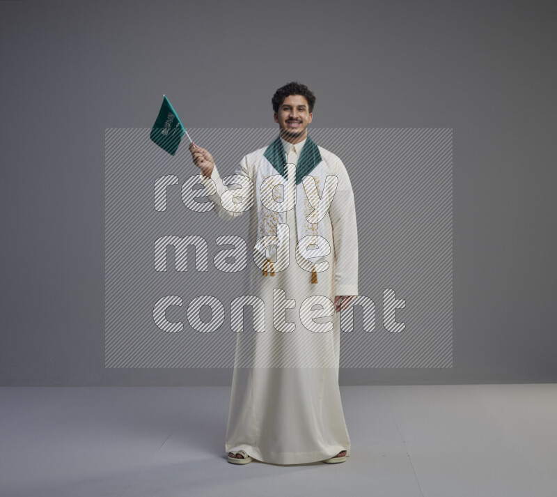 A Saudi man standing wearing thob and saudi flag scarf and holding small saudi flag on gray background