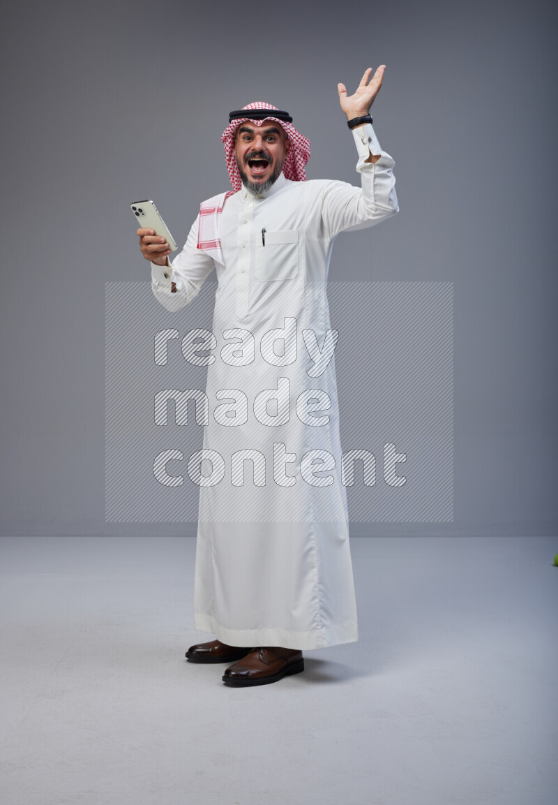 Saudi man Wearing Thob and red Shomag standing texting on phone on Gray background