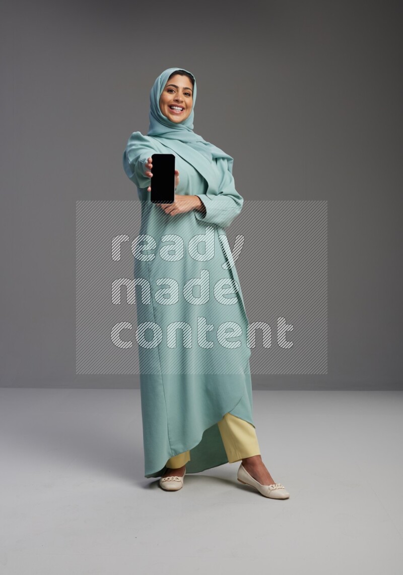 Saudi Woman wearing Abaya standing showing phone to camera on Gray background