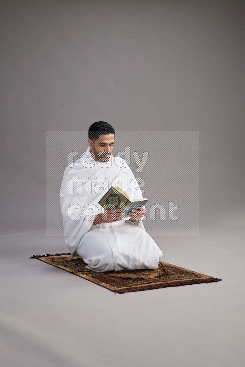 A man wearing Ehram sitting on floor reading quran on gray background