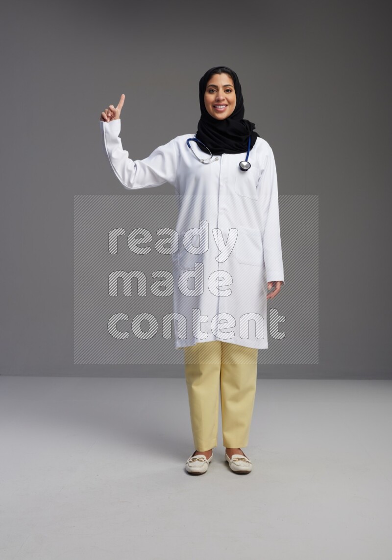 Saudi woman wearing lab coat with stethoscope standing interacting with the camera on Gray background