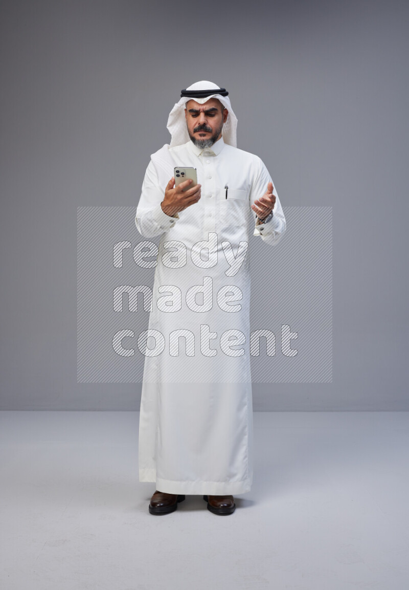 Saudi man Wearing Thob and white Shomag standing texting on phone on Gray background