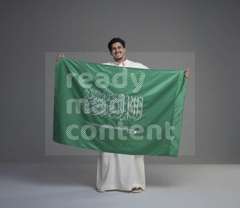 A Saudi man standing wearing thob holding big Saudi flag on gray background