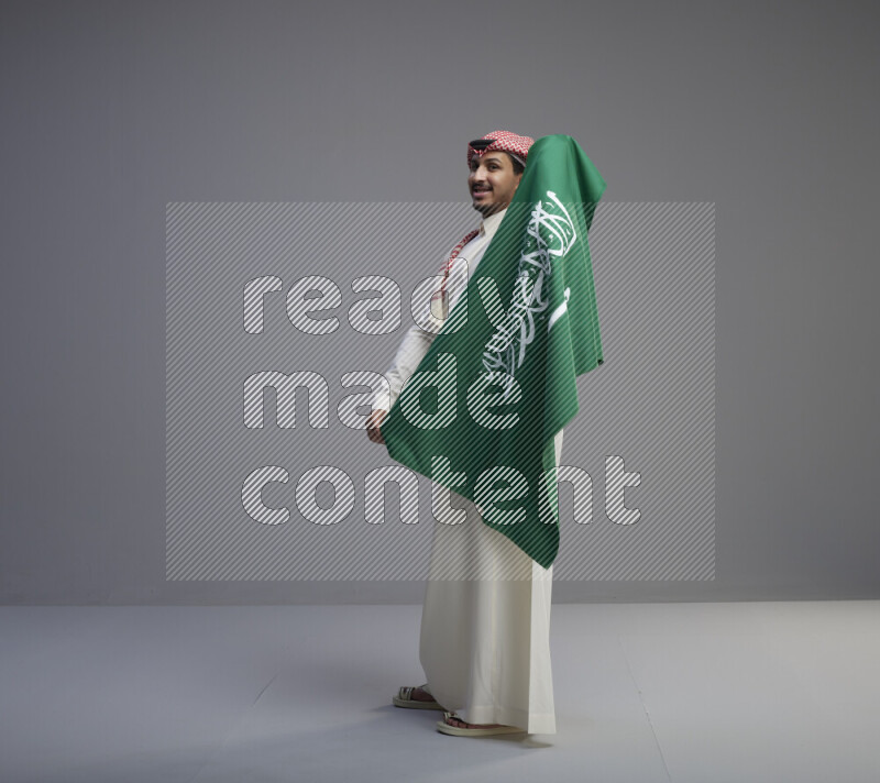 A saudi man standing wearing thob and red shomag holding big saudi flag on gray background
