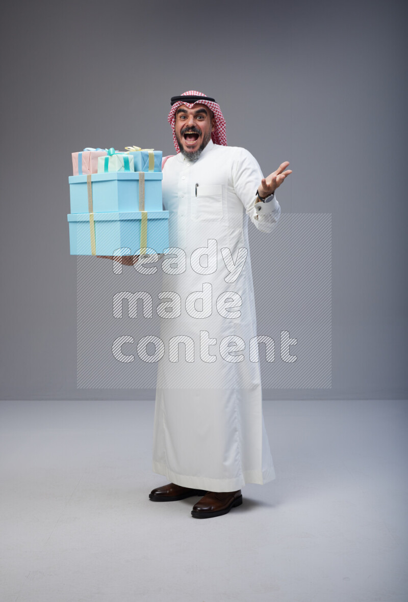 Saudi man Wearing Thob and red Shomag standing holding gift box on Gray background