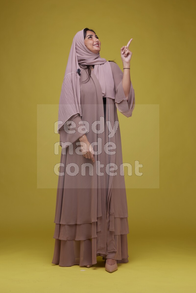 A woman Pointing on a Yellow Background wearing Brown Abaya with Hijab