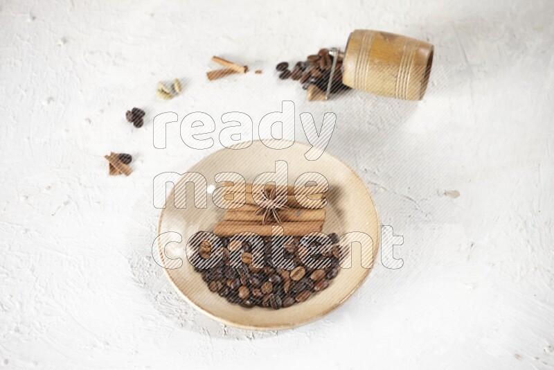 Beige plate full of coffee beans, cinnamon sticks and star anise with a coffee grinder, coffee beans, cinnamon pieces and cardamom next of it on white background
