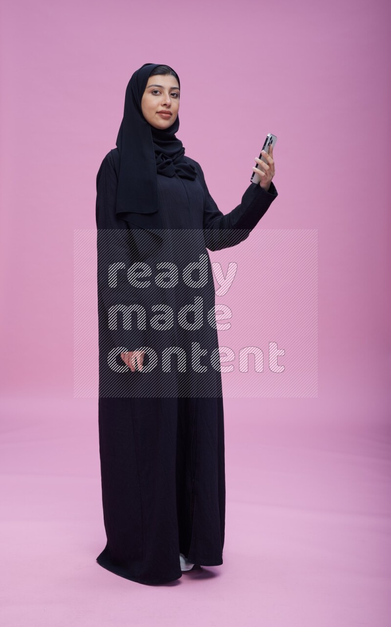 Saudi woman wearing Abaya standing taking selfie on pink background