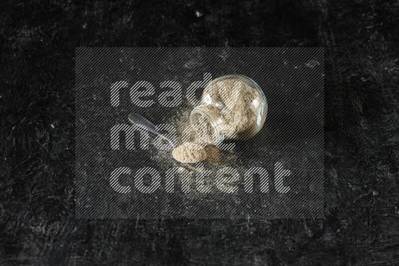 A flipped glass spice jar and metal spoon full of cardamom powder and powder spilled out of it on textured black flooring