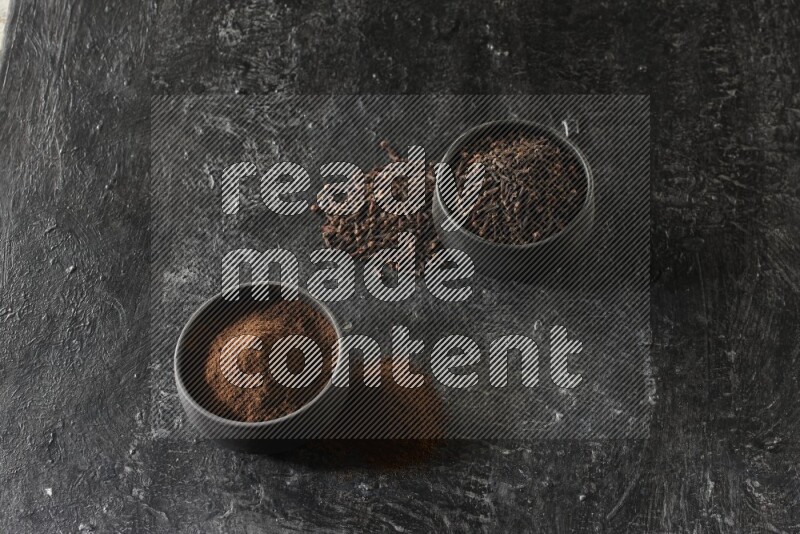 2 Black pottery bowls full of cloves and the other full of cloves powder on textured black flooring