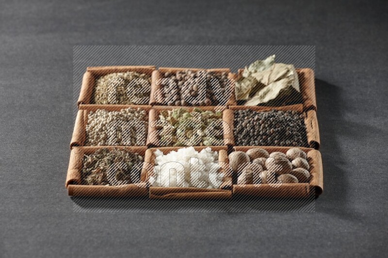 9 squares of cinnamon sticks full of cardamom in the middle surrounded by nutmeg, black pepper, bay laurel leaves, allspice, cumin, white pepper, dried basil and salt on black flooring