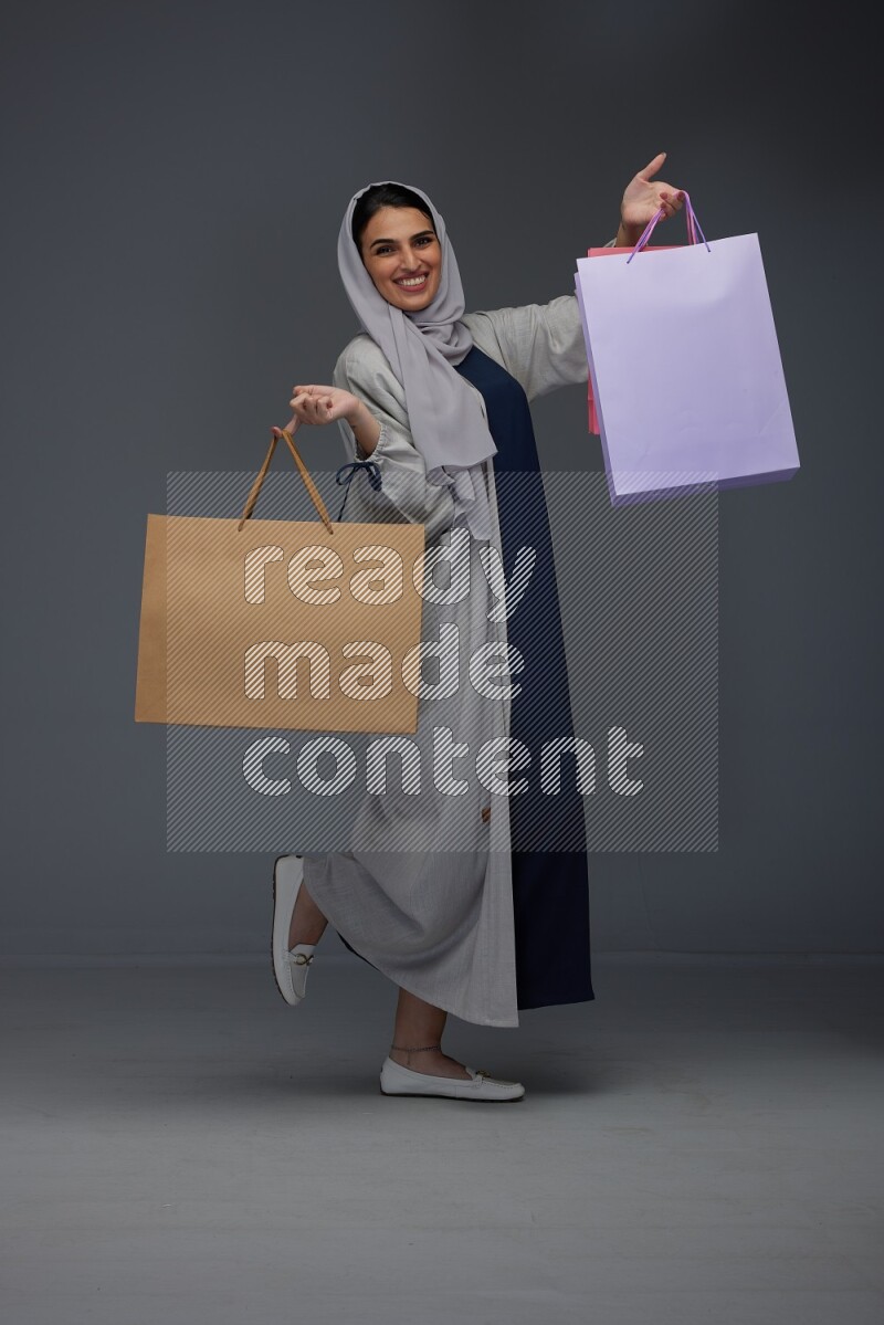 A Saudi woman wearing a light gray Abaya and head scarf standing and holding shopping bags on a grey background