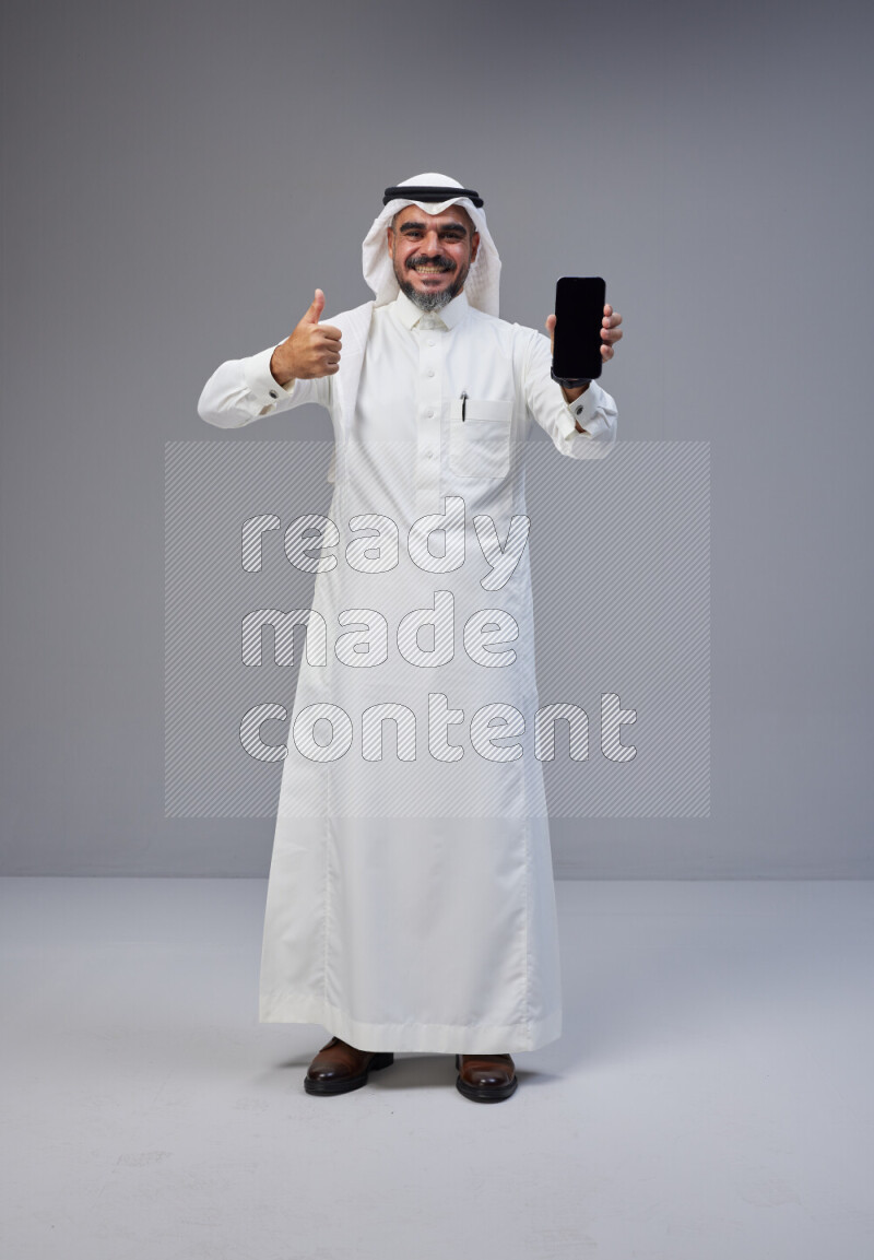 Saudi man Wearing Thob and white Shomag standing showing phone to camera on Gray background