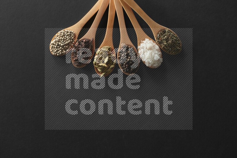 الفلفل الأبيض والقرنفل والهيل والملح والفلفل الأسود وريحان مجفف في معالق خشبية علي خلفية سوداء