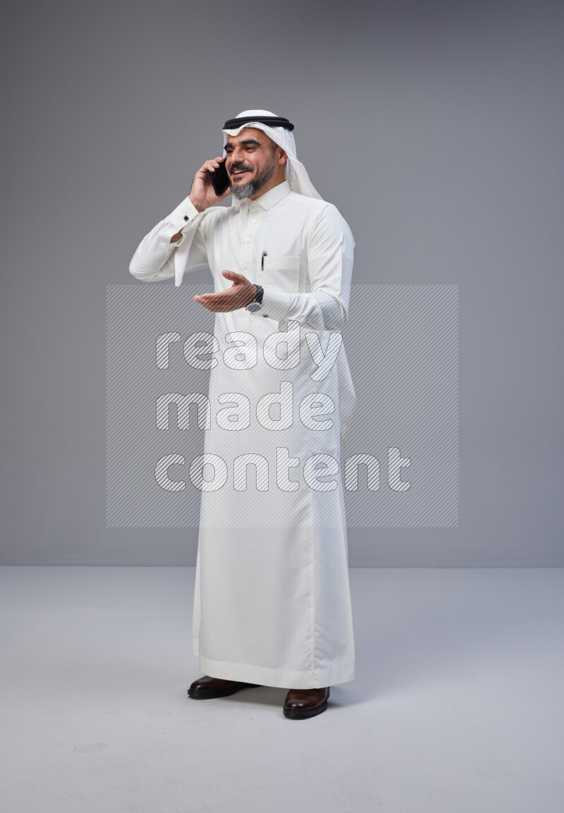 Saudi man Wearing Thob and white Shomag standing talking on phone on Gray background