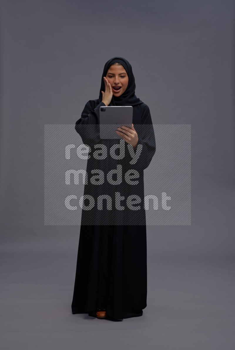 Saudi woman wearing Abaya standing working on tablet on gray background