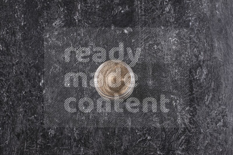 A glass jar full of black pepper powder on a textured black flooring