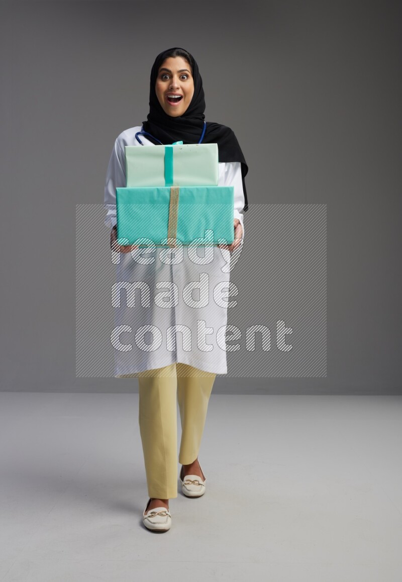 Saudi woman wearing lab coat with stethoscope standing holding gift box on Gray background