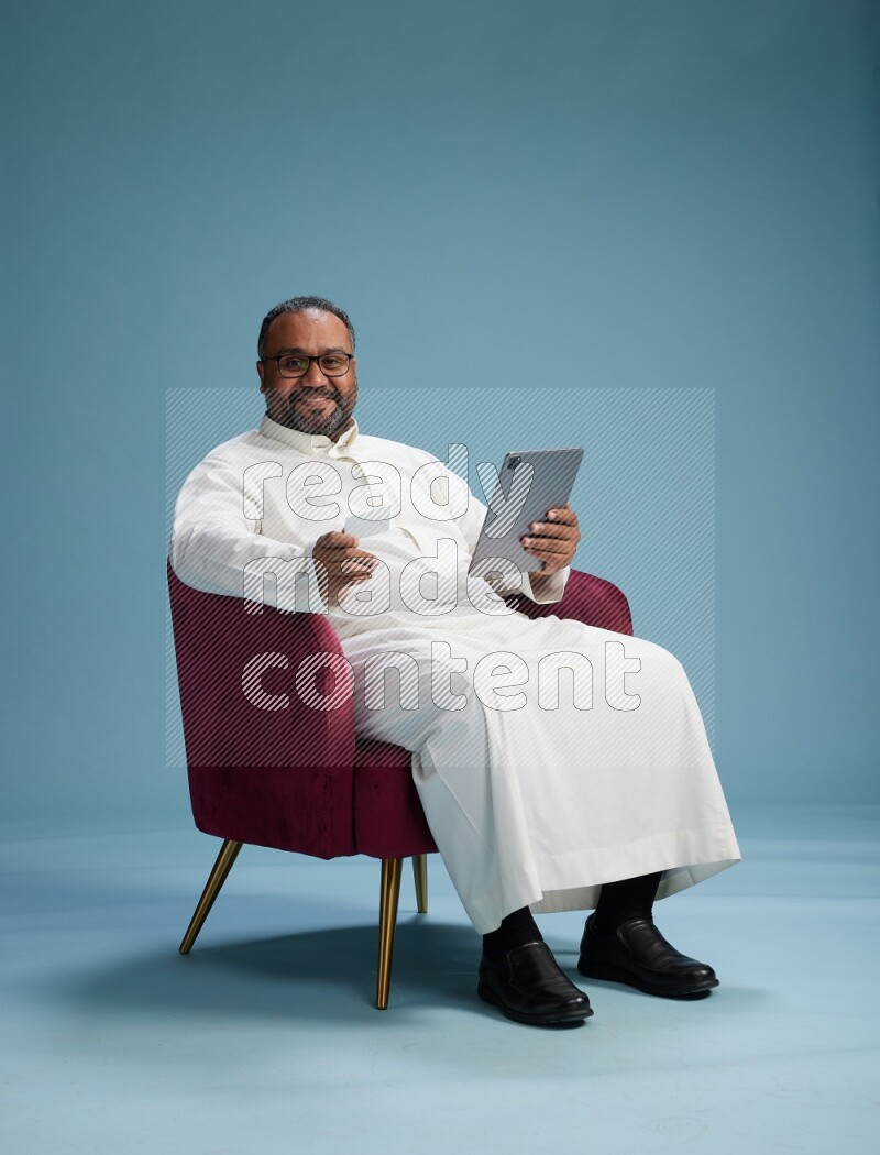 Saudi Man without shimag sitting on chair holding ATM card while working on tablet on blue background