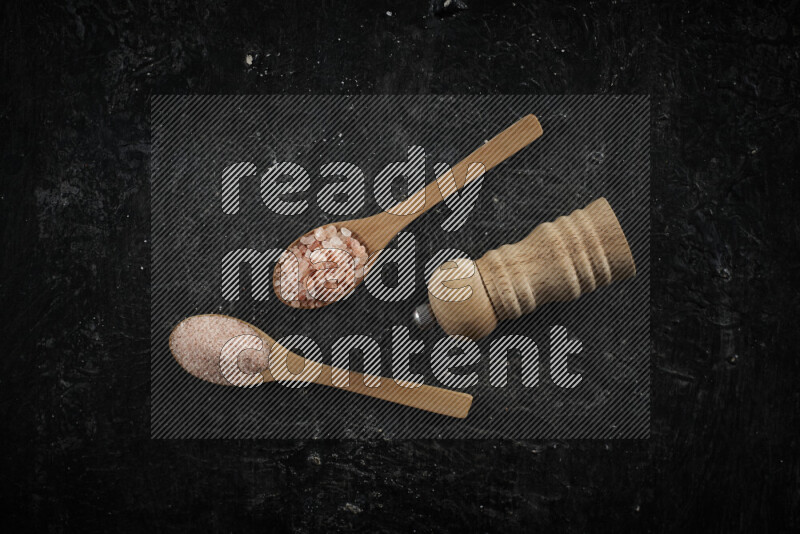 2 wooden spoons filled with fine and coarse salt with wooden grinder beside them on black background