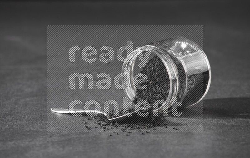 وعاء زجاجي مقلوب ممتلئ ببذور حبة البركة والبذور متناثرة بجانبه مع ملعقة معدنية ممتلئة بالبذور على أرضية سوداء